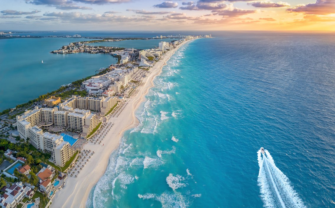 A aerial view shot of Cancun, Mexico with a view of the coast, vibrant clear blue ocean, orange sunset and a boat on the water