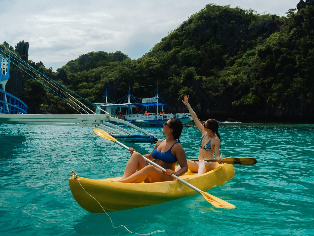 Two female backpackers paddling a yellow kayak in the bright turquoise lagoon waters near a large boat on an El Nido island hopping trip.